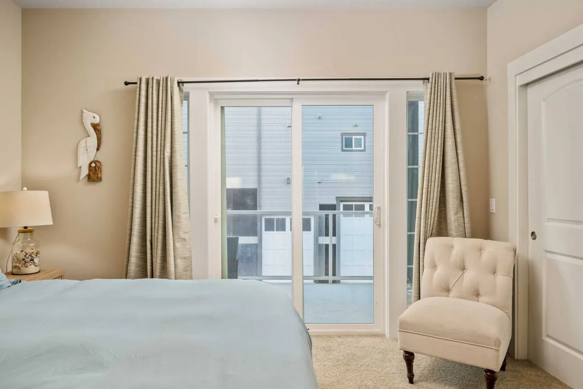 A serene bedroom with light blue bedding, a sliding glass door that likely leads to a deck or balcony, a tufted chair for relaxation, and decorative coastal wall art. The overall mood is soothing and restful.
