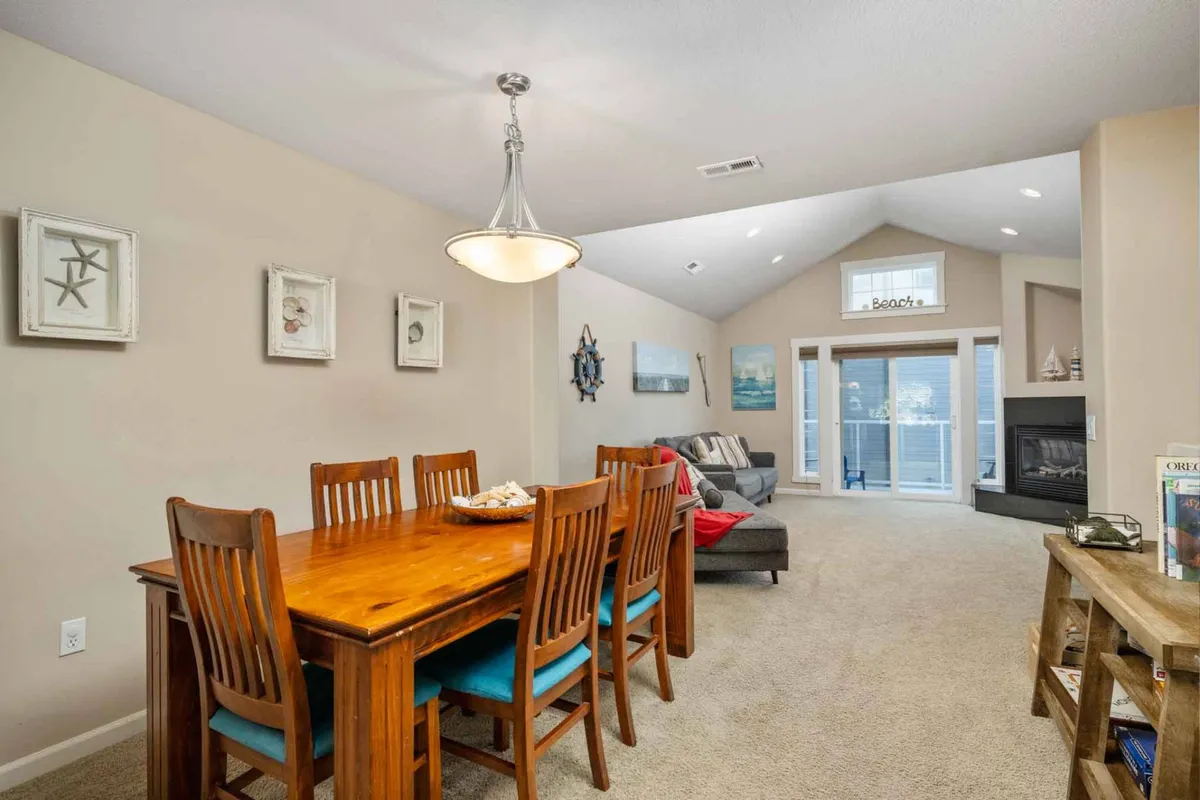 A spacious open-concept interior with a dining table set for a meal, a comfortable sofa, and a cozy fireplace. Sunlight streams in through large windows, accentuating the coastal artwork on the walls. The atmosphere is warm and welcoming, ideal for family dinners, lively game nights, or quiet relaxation by the fire.