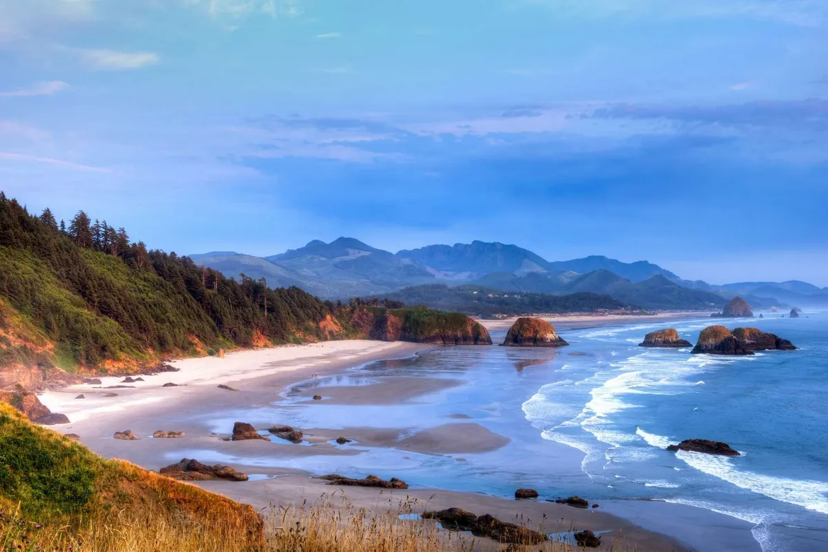 This image captures a sweeping view of the coastline, with rolling ocean waves, striking rock formations, a sandy shoreline, and distant mountains under a tranquil sky. The natural beauty is highlighted by the soft lighting, possibly around sunset, evoking a peaceful and romantic ambiance.