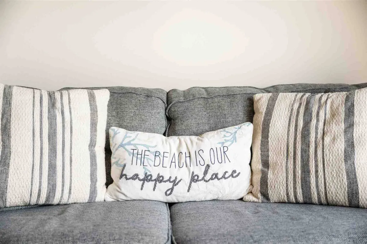 A cheerful, cozy living room with a gray couch decorated with striped and beach-themed pillows, bathed in natural light. The setup invites guests to unwind, socialize, or enjoy a morning coffee while appreciating the subtle nods to the property's beachside location.