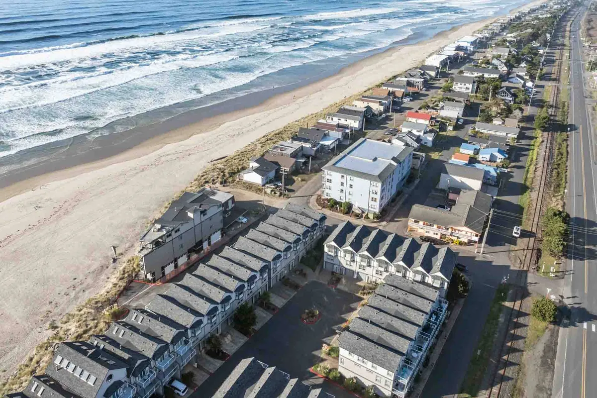 A lively stretch of Rockaway Beach’s coastal road, lined with colorful beach houses and small roadside shops. The shoreline, dotted with ocean waves, creates a seamless connection between town and sea. There are people strolling, exploring shops, and socializing at beachfront cafes, inviting viewers to imagine themselves enjoying the local scene.