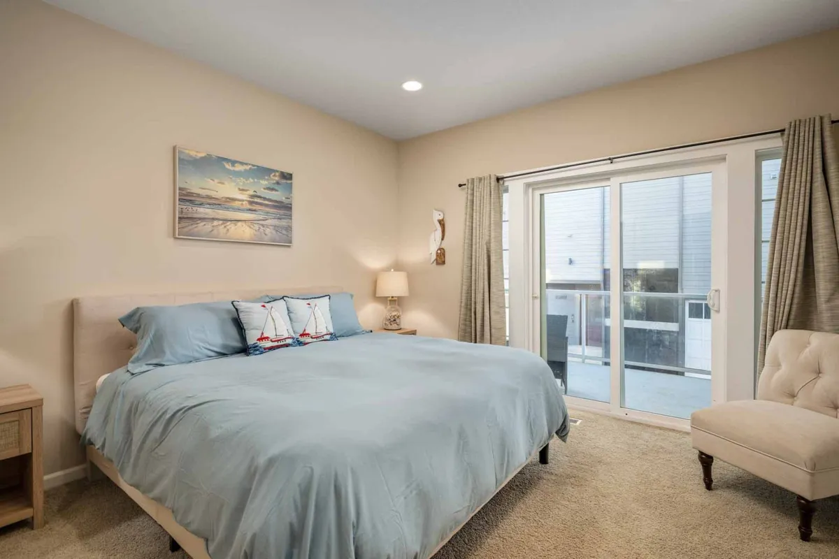 A tranquil bedroom featuring a comfortable bed adorned with soft pillows and plush bedding. A sliding glass door allows natural light to pour in and opens onto an outdoor space, while a lamp and chair add to the cozy, restful ambiance. Subtle coastal artwork on the wall reinforces the beachside retreat theme.