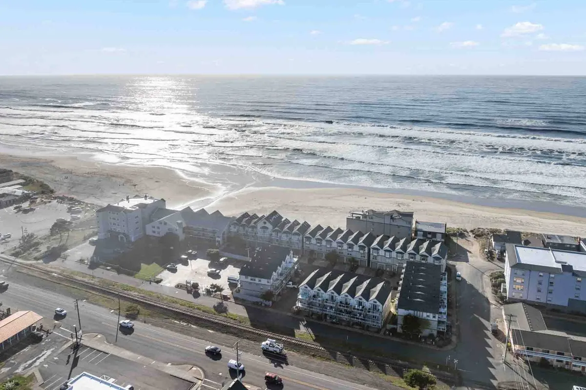 A striking coastal view featuring ocean waves gently rolling onto the beach, with beachfront properties and a coastal road visible in the distance. The scene captures the inviting expanse of sandy shoreline, ideal for dog walks and seaside adventures. The relaxed mood and sense of open space reflect the freedom pets and owners can enjoy together.
