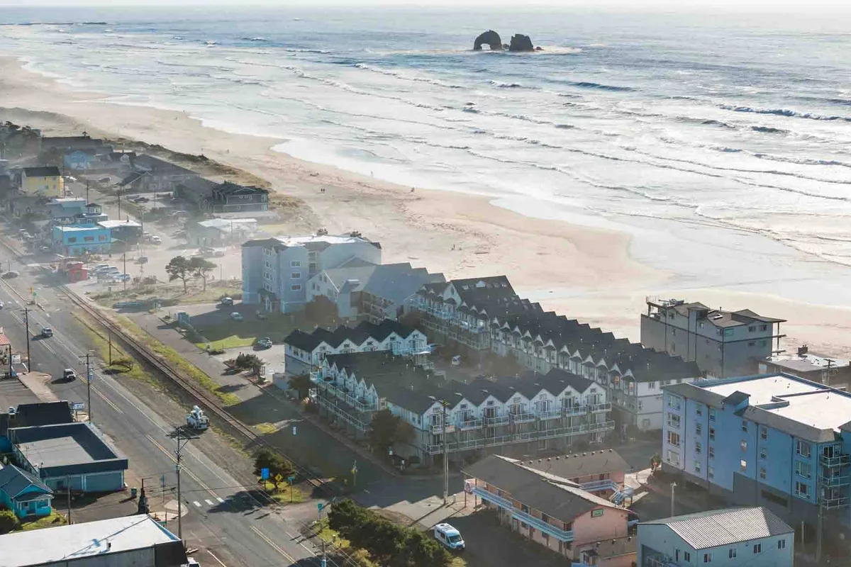 The image showcases the exterior of a stylish condo just steps from the sandy shoreline of Rockaway Beach, with sweeping views of the Pacific Ocean and vibrant coastal buildings. The sun dips low on the horizon, hinting at spectacular sunsets, while the expansive beach invites strolls, beachcombing, and surfside activities. The scene is inviting, blending natural beauty with the ease of beachfront living.