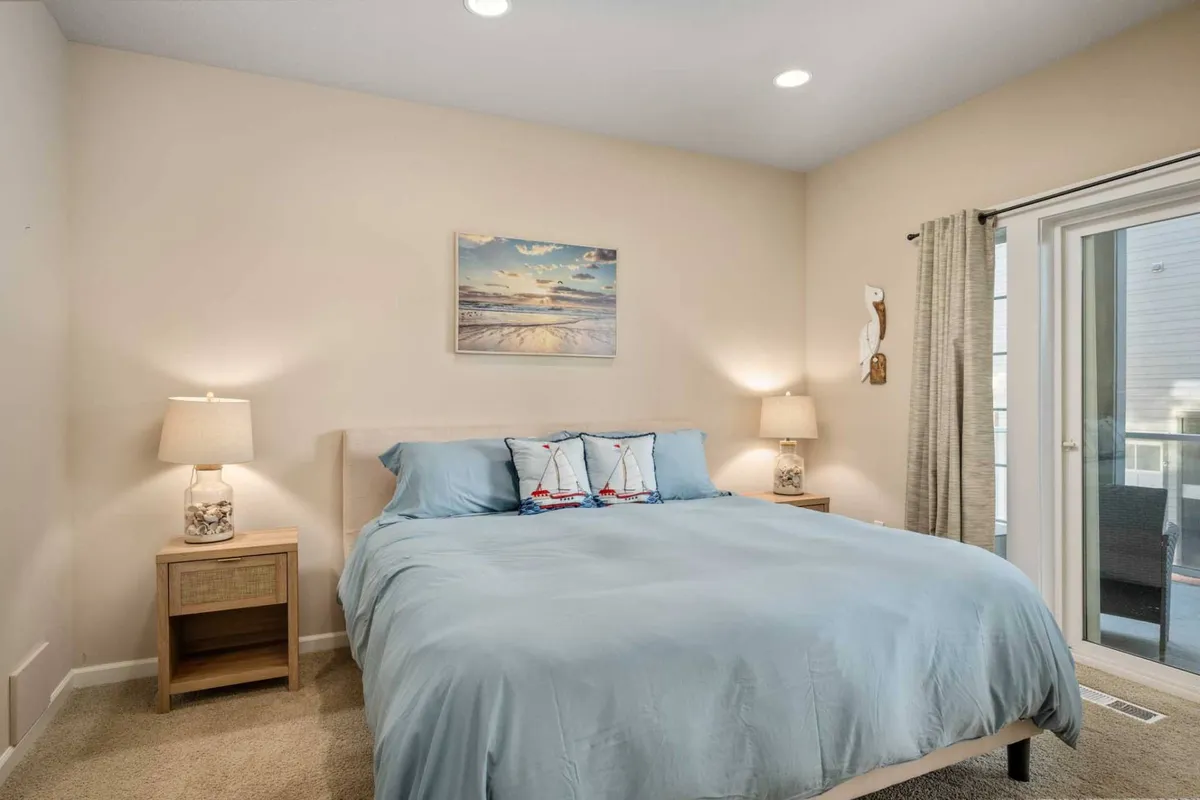A serene bedroom with a bed dressed in light blue bedding, flanked by nightstands and lamps, and adorned with a framed painting of a beach landscape. The space is calm, softly lit, and invites restful sleep or leisurely mornings.